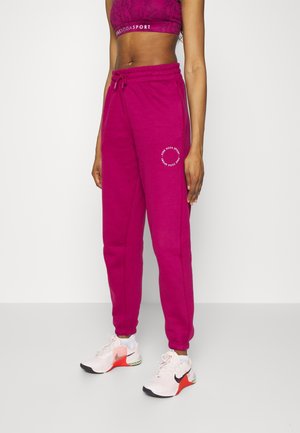 Modèle féminin portant un pantalon de jogging magenta avec des poignets élastiques et un logo blanc, un soutien-gorge de sport rose, et des chaussures de running blanches Nike avec des touches orange.