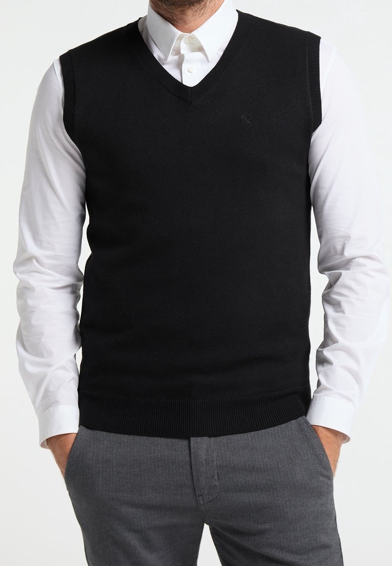 Homme portant un pull sans manches noir à col en V sur une chemise blanche boutonnée avec un pantalon gris, mains dans les poches, fond clair uni.