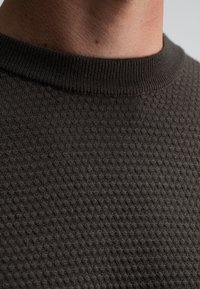 Nahaufnahme des Halses einer Person und eines dunkel strukturierten Strickpullovers mit einem gerippten Rundhalsausschnitt.