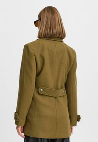 Cappotto in lana verde oliva con vestibilità sartoriale, colletto a punta, polsini con bottoni, dettaglio con cintura sul retro e bottoni in tonalità argentata.