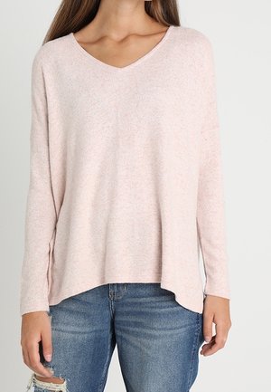 Femme portant un pull à col en V rose clair à manches longues associé à un jean bleu déchiré, visible de la poitrine aux hanches.