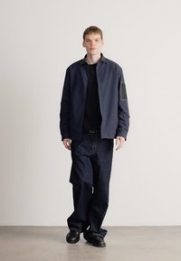 Mornarska jakna z ovratnikom, pod njo črna majica, temne kavbojke in črni čevlji; preprost dizajn z minimalnimi poudarki.