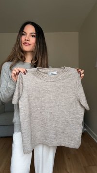 Krémový krátký pletený svetr s kulatým výstřihem a jemnou texturou, který drží osoba oblečená v šedém topu, na neutrálním pozadí.