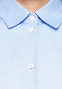 Lyseblå skjorte med krage og teksturert stoff, med tre hvite knapper foran og et rent, enkelt design.