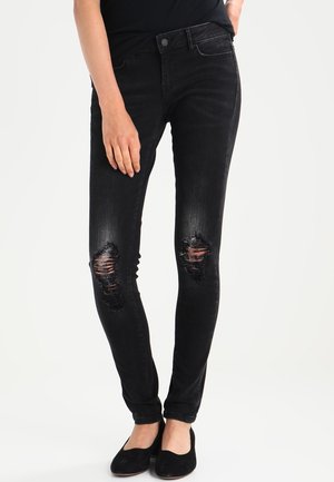 Mujer con jeans pitillo negros desgarrados y zapatos planos negros, de pie con una mano apoyada en el muslo sobre un fondo liso.