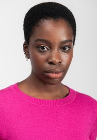 Ung kvinde med kort krøllet hår iført en lys pink sweater, der kigger direkte ind i kameraet med et neutralt udtryk.