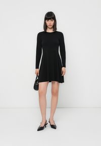 Black long-sleeve dress with a flared skirt, made of smooth fabric. Model holds a small black handbag and wears pointed black heels.