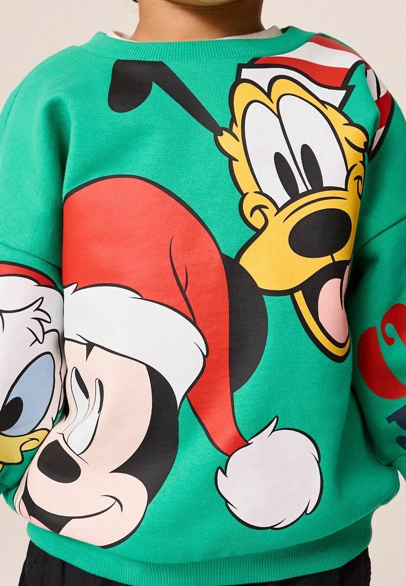Enfant portant un sweat-shirt vert avec de grands visages de dessin animé de Mickey, Donald et Pluto, tous portant des chapeaux de Noël rouges et blancs.