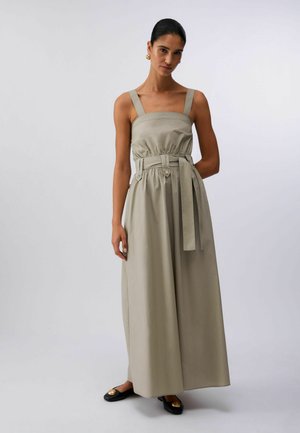 Donna che indossa un abito lungo beige senza maniche con spalline larghe, una cintura in vita e ballerine nere con decorazioni dorate, che posa davanti a uno sfondo semplice.