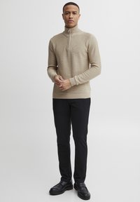 Beige bordás kötött pulóver magas cipzáras gallérral, fekete slim-fit nadrággal és fekete cipővel párosítva, kiemelve a textúrát és a dizájnt.