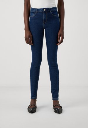 Skinny denim jeans in dark blue, high-waisted design, featuring five pockets and subtle stitching. Worn with black patterned flats.