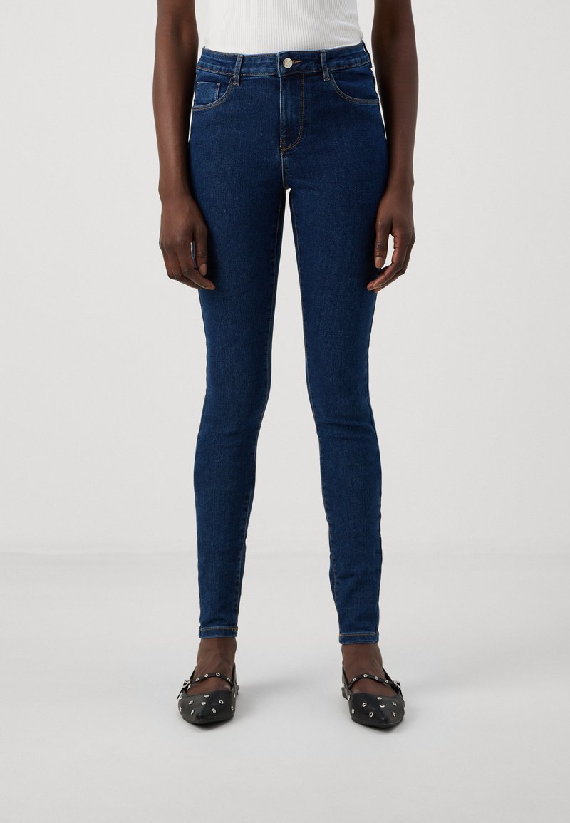 Λεπτά τζιν παντελόνια σε σκούρο μπλε, ψηλόμεσο σχέδιο, με πέντε τσέπες και διακριτή ραφή. Φορεμένα με μαύρες φλατ παπούτσια με σχέδιο.