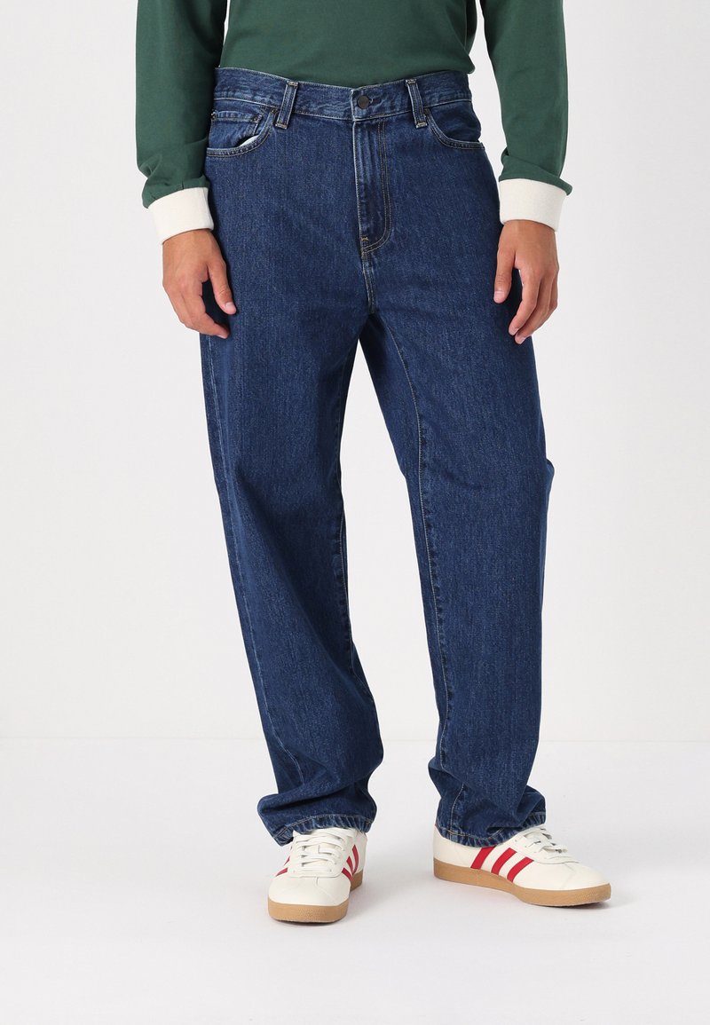 Osoba oblečená v volných tmavě modrých džínách, bílých teniskách s červenými pruhy a zelené košili s dlouhým rukávem a krémovými manžetami, stojí na bílé podlaze.