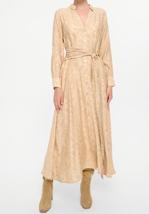 Langes, beiges Kleid mit strukturiertem Wellenmuster, Knopfleiste, Kragen und tailliertem Gürtel. Kombiniert mit taupefarbenen kniehohen Stiefeln.