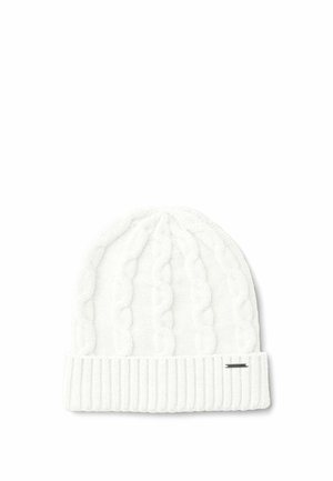 Femme portant un bonnet en laine blanc, des gants blancs, une veste matelassée bleu marine et une chemise rayée, souriante et regardant vers le bas en ajustant sa veste.