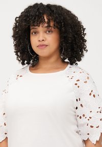 A woman with curly hair wears a white top with lace sleeves, looking directly at the viewer with a neutral expression.