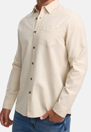 Homme portant une chemise crème texturée à manches longues avec poche poitrine et jeans bleus, main dans la poche, sur fond uni.