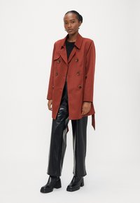 Balonar plašč rjave rjave barve z dvovrstnim zapenjanjem, rjavimi gumbi in stranskimi trakovi, kombiniran s sijočimi črnimi širokimi hlačami in masivnimi črnimi škornji.