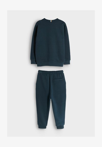 Sötétkék pulóver és melegítőnadrág szett. A pulóver bordázott mandzsettákkal rendelkezik; a nadrág rugalmas derékkal és hátsó zsebbel rendelkezik. Puha szövettextúra.