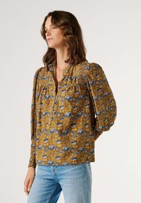 Florale Bluse in Senfgelb mit blauen und weißen Mustern. Verfügt über eine Knopfleiste, Smokdetail und lange, aufgeplusterte Ärmel.