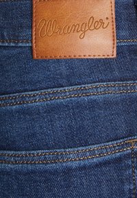 Tela de mezclilla de un azul oscuro con líneas de costura visibles, que cuenta con una etiqueta de cuero marrón con el texto "Wrangler" en relieve.