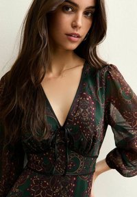 Jurkje met lange mouwen in donker groen met een paisley-patroon in bordeauxrood en goud. Heeft een diepe V-hals en een stoffen ceintuur in de taille.