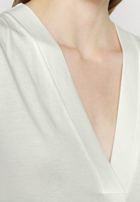 Haut blanc à col en V avec une texture douce et lisse. Présente un décolleté profond et des manches courtes, visible depuis la zone de la clavicule.