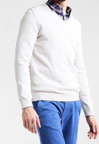 Homme portant un pull blanc à col en V par-dessus une chemise à carreaux, associé à un pantalon bleu, debout devant un fond blanc uni.
