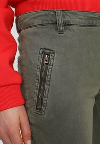 Olivgröna byxor med en dragkedjeficka; tyget har en slät yta. Röd tröja synlig överst med ribbad manschettdetalj.