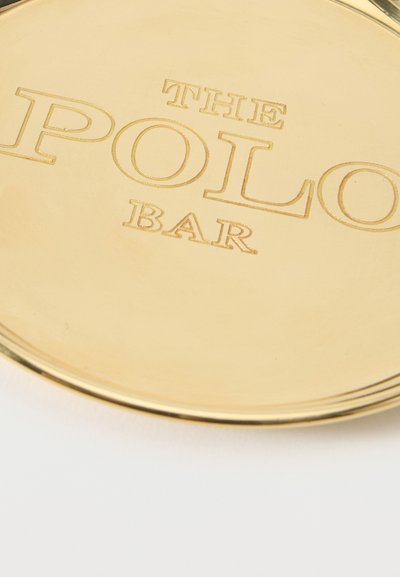 Κοντινή λήψη στρογγυλής δίσκου σε χρυσαφί χρώμα με χαραγμένο το κείμενο "THE POLO BAR" με κεφαλαία γράμματα.
