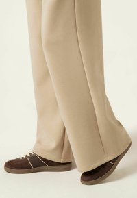Beige vida byxor med en slät textur, ihop med mörkbruna sneakers med ljusa detaljer och beige snören, sedd från vristen och ner.