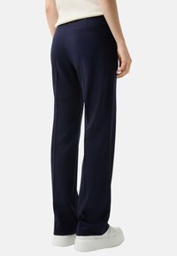 Pantalon ajusté bleu marine en tissu lisse et léger. Comprend une poche arrière et une coupe droite. Porté avec des baskets blanches.