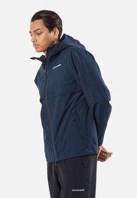 Mandlig model iført en marineblå hættetrøje og sorte bukser, stående med hænderne bag ryggen og vendt mod venstre på en hvid baggrund.