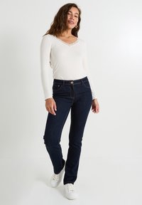 Top de manga larga color crema con cuello de encaje, combinado con jeans de mezclilla azul oscuro y zapatillas blancas, de pie frente a un fondo claro.