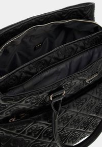 Borsa a tote nera trapuntata con un'esterno liscio, doppie tasche con zip e un logo distintivo. Interno foderato in tessuto dai toni scuri.