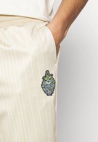 Hand i fickan på beiga randiga byxor med broderad Fila Golf-logga som har korslagda golfklubbor och en ananas.