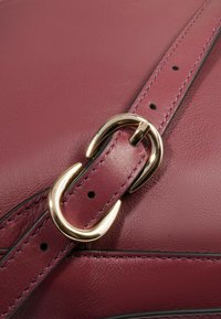 Bolso de cuero burdeos con textura suave, que cuenta con una hebilla metálica dorada y una correa con costuras visibles y orificios de ajuste.