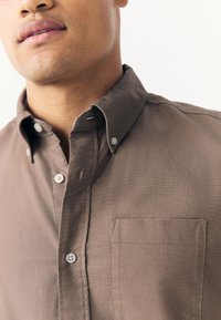 Camisa marrón abotonada con cuello, hecha de tela tejida. Cuenta con un bolsillo en el pecho y botones blancos, mostrando una textura suave.