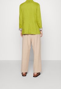 Persona che indossa una giacca a maniche lunghe verde lime, pantaloni beige a gamba larga e sandali marroni, in piedi su uno sfondo bianco semplice.