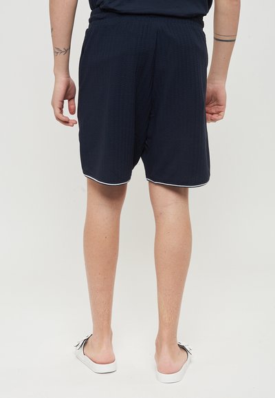 Costumi da bagno blu navy con un motivo testurizzato, vita elastica e bordo bianco all'orlo. Indossati con scarpe bianche senza lacci.