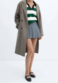 Cappotto grigio doppiopetto sopra un maglione a righe verde e bianche, abbinato a una gonna plissettata grigia e a delle ballerine nere con dettagli a cinturino.
