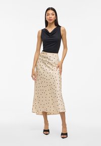 Črna bluzica s kapuco v obliki cowl, kombinirana s svetlo bež satensko krilo z črnimi pikami. Obleka ima gladko teksturo in sega pod kolena.