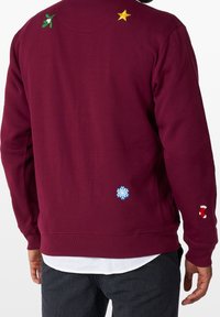 Vinröd sweatshirt med broderade julmotiv: mistel, stjärna, snöflinga och godistrand. Slät textur och avslappnad passform.