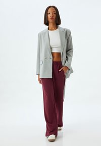 Lichtgrijze dubbelknoop blazer over een witte crop top, gecombineerd met bordeaux kleurige wijde sweatpants met grijze zijpanden en beige schoenen.