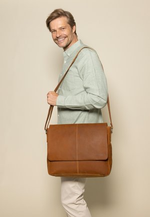 Lächelnder Mann mit hellgrünem Hemd und beigen Hosen trägt eine große braune Lederschultertasche vor beigem Hintergrund.