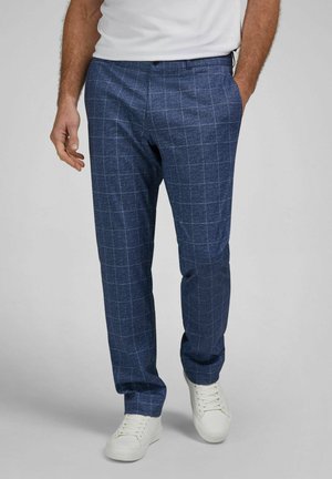 Mann trägt blaue karierte Hose mit weißen Turnschuhen und weißem Hemd, steht mit einer Hand in der Tasche vor schlichthintergrund.