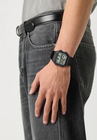 Montre numérique avec un bracelet en résine noire, un cadran rectangulaire, un cadran gris avec écran LCD, plusieurs cadrans et des accents de contraste. Portée sur une main.