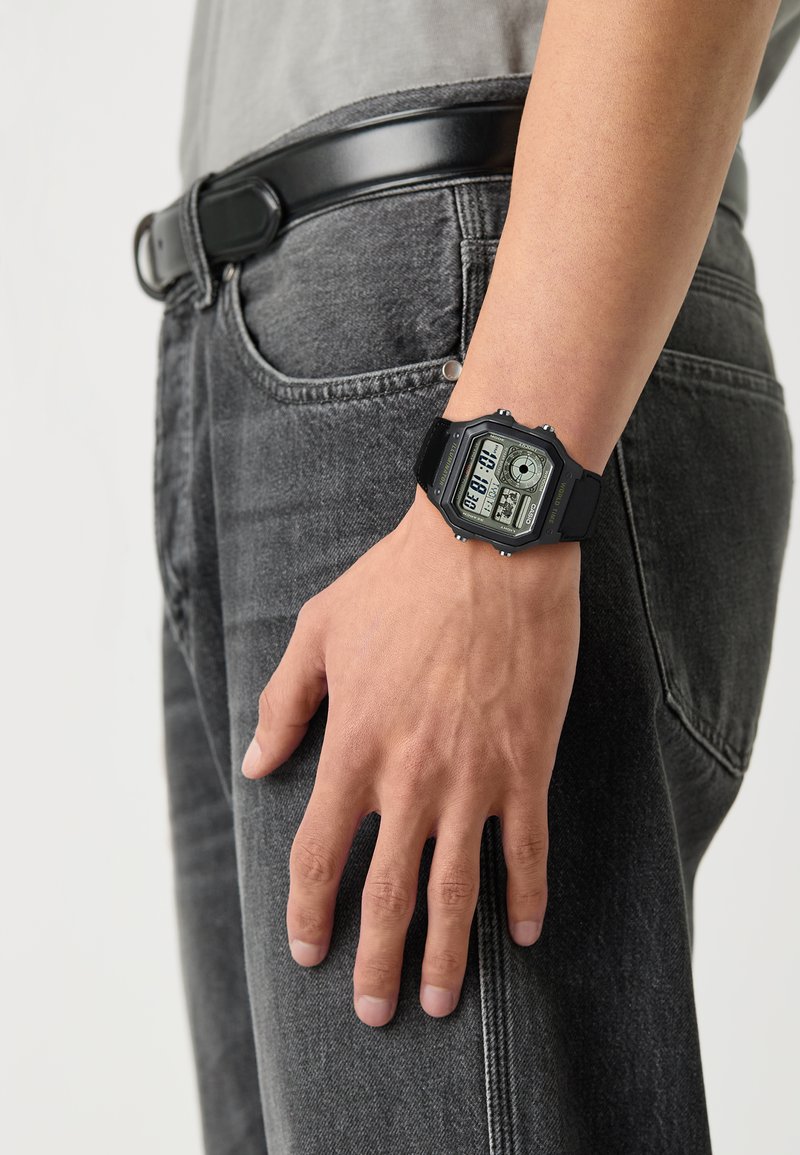 Montre numérique avec un bracelet en résine noire, un cadran rectangulaire, un cadran gris avec écran LCD, plusieurs cadrans et des accents de contraste. Portée sur une main.