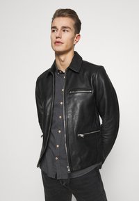 Chaqueta de cuero negra con un collar puntiagudo, dos bolsillos de pecho con cremallera y dos bolsillos laterales con cremallera, llevada sobre una camisa oscura de botones.