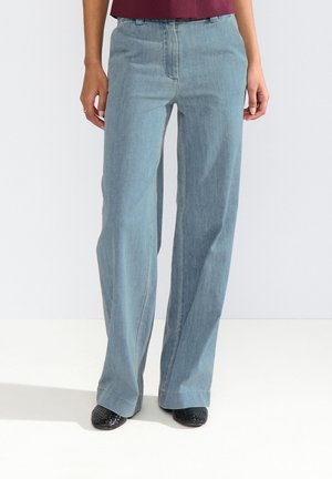 Person trägt hellblaue Jeans mit weitem Bein und schwarze strukturierte Schuhe, steht vor einem einfachen hellen Hintergrund.